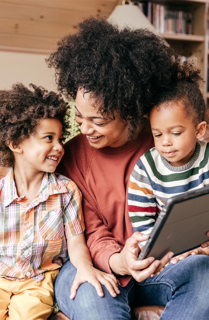 A Black woman smiles and holds a tablet as two Black children sit in her lap, bookcase in the background.