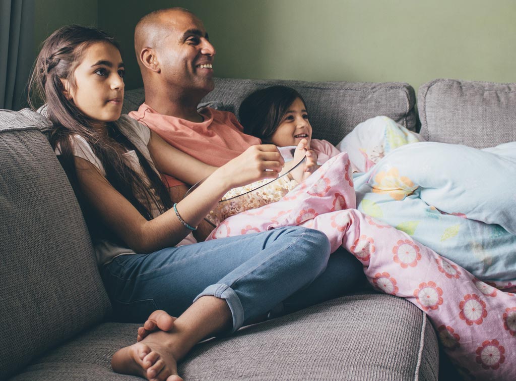 A smiling man with brown skin sits on a couch between two girls with brown skin, as they all look off to the right.